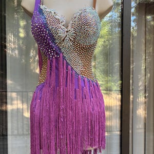 May include: A purple dance dress featuring a sweetheart neckline, thin straps, and an asymmetrical hemline. The bodice is adorned with dense, iridescent crystal embellishments and white lace accents, while the skirt consists of long purple fringe.