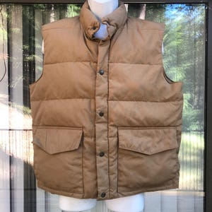 May include: A tan puffer vest with a high collar and two front pockets. The vest has a button-up closure and is displayed on a white mannequin. The vest is made of a quilted material.