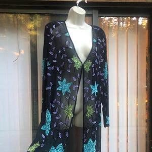 May include: Black open-front longline cardigan adorned with turquoise, green, and purple sequined floral and leaf designs. The sheer fabric drapes elegantly, creating a flowing silhouette. This embellished cardigan is a statement piece.