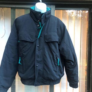May include: Black winter jacket with a teal-colored interior lining and accents. The jacket has a high collar, two front pockets, and two side pockets with teal zippers. The jacket is buttoned up and hanging on a white mannequin.