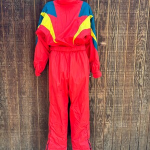 May include: A vintage red ski suit with teal and yellow detailing. The one-piece suit has a high collar, long sleeves, and zipped pockets. The suit is hanging on a metal hanger against a wooden backdrop.