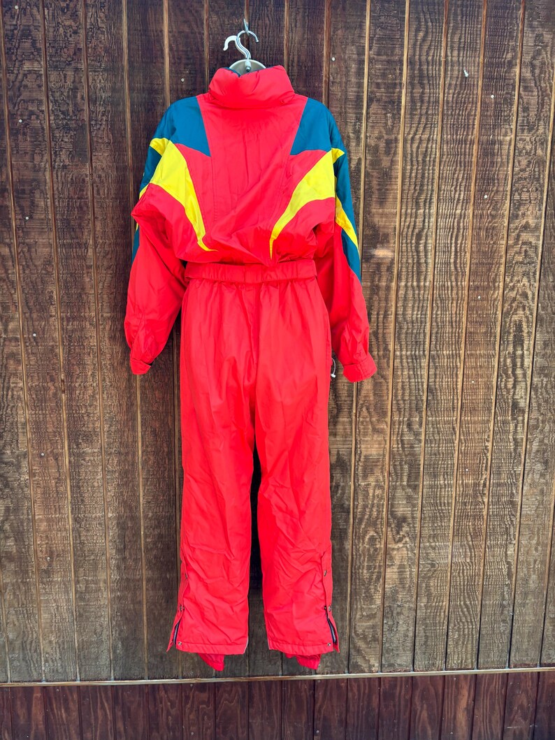 May include: A vintage red ski suit with teal and yellow detailing. The one-piece suit has a high collar, long sleeves, and zipped pockets. The suit is hanging on a metal hanger against a wooden backdrop.