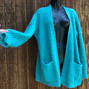 May include: A teal blue open-front cardigan with long sleeves and two pockets. The cardigan is made of a soft, fuzzy material.