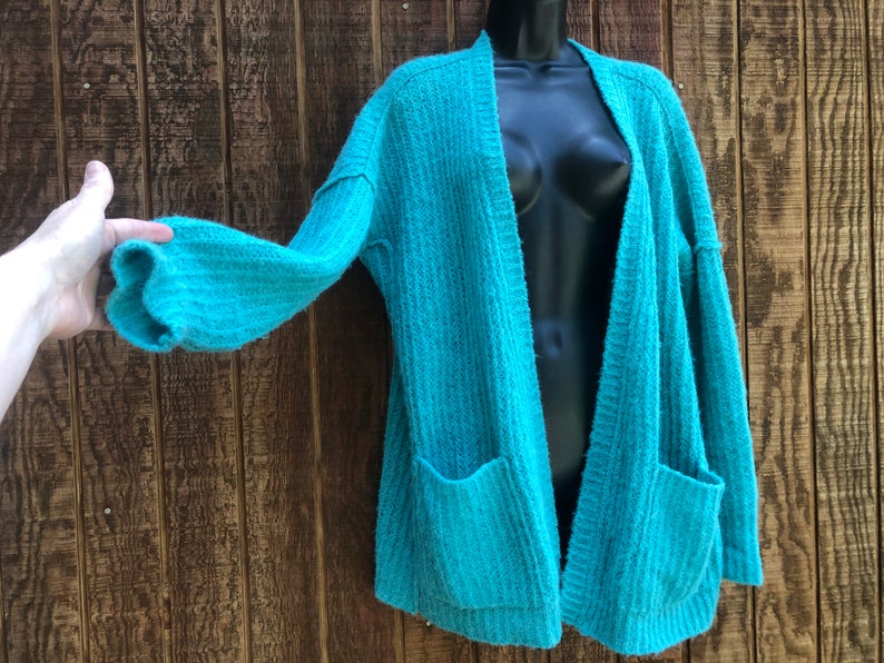 May include: A teal blue open-front cardigan with long sleeves and two pockets. The cardigan is made of a soft, fuzzy material.