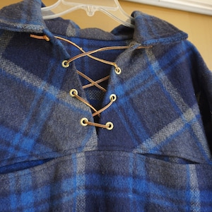 May include: A blue and gray plaid wool coat with a collared neckline and a laced closure. The coat has a pocket and is hanging on a hanger. The coat is made of a soft, warm material.