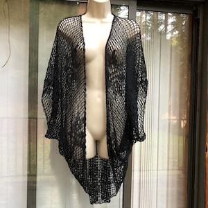 May include: Black open-weave cardigan displayed on a mannequin. The cardigan has a loose, flowing design with a wide, open front and long sleeves. The knit pattern creates a net-like appearance. The garment is made of a soft, lightweight material.