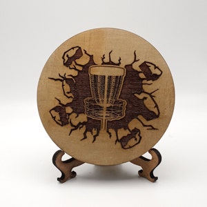 May include: A wooden disc coaster with a laser-engraved design of a disc golf disc breaking through the surface. The coaster is on a small wooden stand.