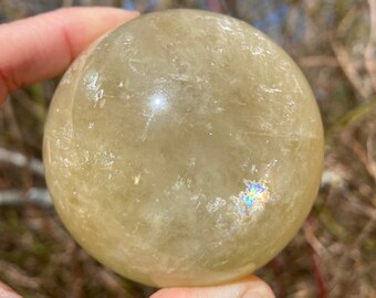 May include: A large, smooth, yellow crystal ball with iridescent rainbow flashes. The ball is being held in a hand.