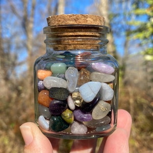 Puede incluir: Un tarro de cristal transparente lleno de una variedad de piedras preciosas pulidas de colores. El tarro tiene un tapón de corcho y se sostiene contra un fondo natural borroso. Las piedras preciosas incluyen tonos de púrpura, verde, naranja y transparente.