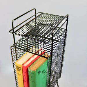 May include: A black metal magazine rack with a perforated design, holding three vintage books with colorful covers. The rack has a top shelf and a lower compartment for storage, with a minimalist design.