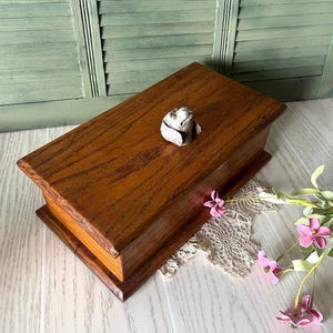 May include: A rectangular wooden box with a medium brown wood grain finish and a hinged lid. A small, sculpted, off-white ceramic dog figurine is attached to the center of the lid, serving as a decorative handle for opening the box.