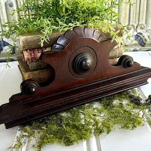 May include: A dark brown wooden shelf bracket featuring a decorative scalloped top edge, a central circular medallion, and two smaller side finials. The piece has a horizontal base and a flat back, showcasing a polished wood grain finish throughout.