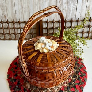 May include: A round, brown woven basket with a handle. The lid is adorned with a small porcelain figurine surrounded by various buttons. The basket sits on a red, brown, and green braided mat.