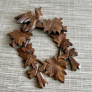 May include: A carved wooden wreath featuring a bird and maple leaves. The bird has detailed wings and a red eye. The leaves are intricately carved with visible veins. The wood has a rich, dark brown color. The wreath is a decorative item.