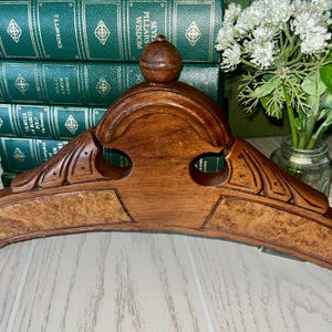 May include: A curved wooden architectural pediment featuring a central finial, carved leaf motifs on the sides, and two inlaid panels with a burl wood grain pattern. The dark brown wood has a polished finish and a symmetrical, arched structure.