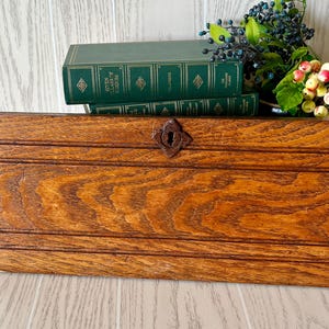 May include: A rectangular wooden box with a prominent brown wood grain pattern and a dark, diamond-shaped metal keyhole escutcheon centered on the front panel. The box features horizontal grooved lines across the top and bottom sections.