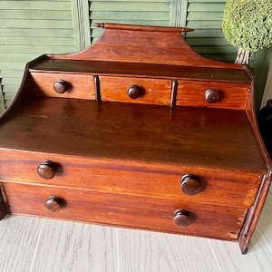 May include: A wooden desktop organizer featuring a dark brown finish with a curved top backboard. It includes three small upper drawers and two larger lower drawers, each equipped with a round wooden knob for opening and closing the compartments.