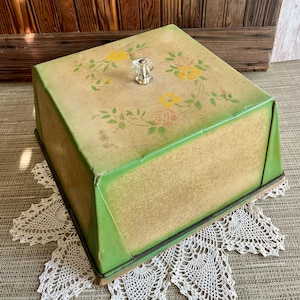 May include: Vintage square cake carrier with a light green exterior and floral design. The lid features a clear glass knob and a painted pattern of yellow and pink flowers with green leaves. The carrier sits on a wooden base.