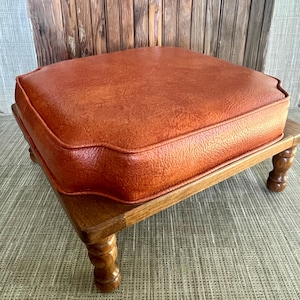 May include: A vintage footstool with a square, orange cushion and a wooden frame. The cushion has a textured surface and a contrasting trim. The legs are intricately carved, adding a touch of elegance to the piece. The footstool is a warm, inviting accent.