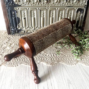 May include: A vintage rolling pin with a brown tweed-like fabric roller and dark wooden handles. The rolling pin is supported by two legs and is designed for kitchen use. The overall aesthetic is rustic and antique.