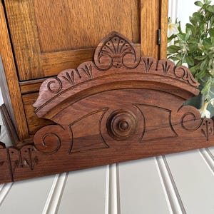 May include: A dark brown wooden architectural pediment featuring intricate carved floral and fan-like motifs. The piece has a central circular medallion and a curved top edge with symmetrical engravings, showcasing a rich wood grain texture.