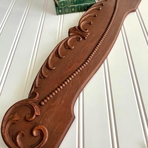 May include: A long, rectangular wooden architectural element with a medium brown finish. The piece features carved scrollwork at both ends, a central carved leaf motif, and a raised beaded trim detail running diagonally across the surface.