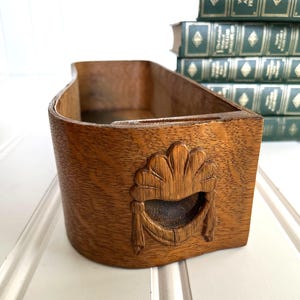 May include: A curved wooden storage tray featuring a prominent carved shell motif on the front panel. The tray has a natural wood grain finish and an open top design, with a smooth interior surface and a sturdy, rounded exterior structure.