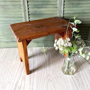 May include: A small, rectangular wooden stool with a weathered brown finish and four sturdy legs. Beside the stool sits a clear glass vase containing a bouquet of white flowers with green leaves, featuring small, delicate, clustered white blossoms.