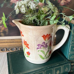 May include: A cream-colored ceramic pitcher with a visible crackle glaze finish, decorated with vibrant red and purple tulip illustrations. The pitcher features a curved handle, a pouring spout, and contains a bouquet of green and white artificial flowers.