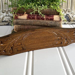 May include: A dark brown wooden decorative piece featuring a central oval panel surrounded by carved leaf and geometric patterns. The object has a horizontal orientation with a smooth, polished finish and detailed relief carvings on both sides.