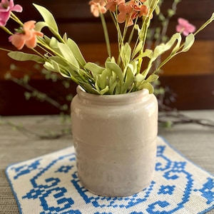 May include: A cylindrical, off-white ceramic crock holds a bouquet of artificial peach and pink flowers with green leaves. The crock sits on a square, white fabric mat featuring a blue, geometric, cross-stitch style pattern.