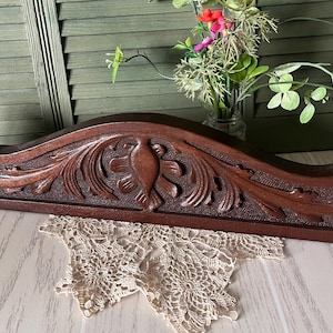 May include: A dark brown wooden architectural pediment featuring an intricate hand-carved relief design of central floral motifs flanked by scrolling acanthus leaves. The piece has a smooth, polished finish and a curved top edge with decorative side accents.