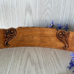 May include: A curved, rectangular wooden block with a medium brown finish, featuring dark, carved scrollwork patterns on the left and right sides. The top edge of the wooden piece contains a series of small, evenly spaced circular holes.