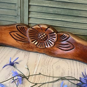 May include: A curved, medium-brown wooden headboard or decorative panel featuring a central carved sunflower motif with detailed petals and a textured center, flanked by smooth, rounded end posts with a polished, glossy finish.