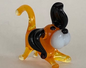 May include: A small glass figurine of a dog with a black and orange body and black ears. The dog has a white face and a curled tail.