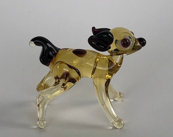 May include: A small glass figurine of a dog. The dog is yellow with brown spots and has black ears. The dog is standing on all four legs and has its head tilted to the side.