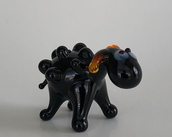 May include: A small, black glass figurine of a four-legged creature with a rounded body, multiple raised bumps along its back, and a head featuring two light blue eyes and curved, amber-colored horns extending from the sides of its head.