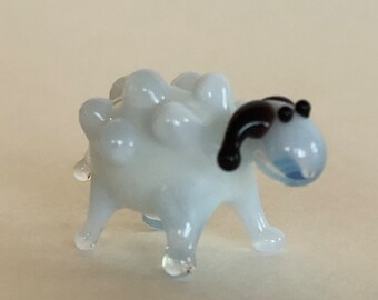 May include: A small white glass sheep figurine with black eyes and a black ear. The sheep has a bumpy, textured body.