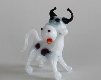 May include: A small glass figurine of a cow with a white body, black spots, and black curved horns. The cow has a white snout with a small red dot, dark eyes, and a thin tail. The glass is smooth and glossy, standing on four legs.