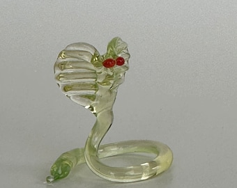 May include: A small glass cobra figurine with a green body and red eyes. The cobra is coiled in a defensive posture.