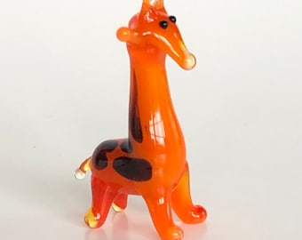 May include: A small glass giraffe figurine with an orange body and black spots. The giraffe has a long neck and a small head with black eyes.