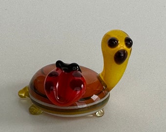 May include: A small glass turtle figurine with a yellow head and a brown, white, and yellow striped shell. A red ladybug with black spots is perched on the turtle's back.