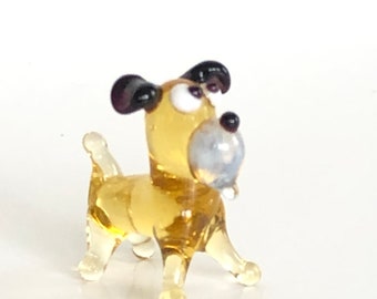 May include: A small glass figurine of a dog. The dog is yellow with black ears and a white face. It has a small, white ball in its mouth.