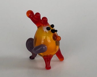 May include: A small glass rooster figurine with a yellow body, orange comb and wattle, black eyes, and purple legs.