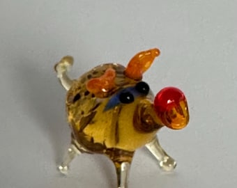 May include: A miniature glass figurine of a pig with a translucent amber-colored body, clear glass legs, and a curled tail. The pig features two orange ears, small black eyes with blue accents, and a prominent red snout at the end of its nose.