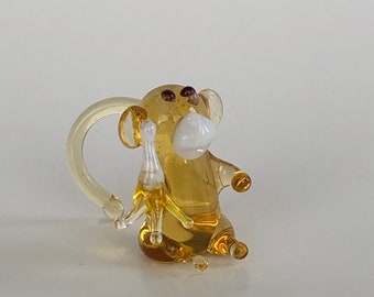 May include: A small, translucent amber-colored glass monkey figurine featuring a white muzzle, dark brown eyes, and a curved tail. The monkey is depicted in a seated position, holding a small, pale yellow banana-shaped object in one hand.