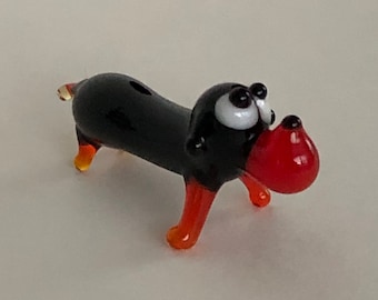 May include: A small glass figurine of a black dachshund with red and orange legs and a red nose. The figurine has white eyes and black pupils.