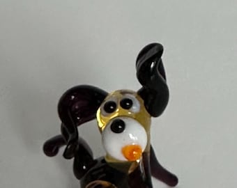 May include: A small glass figurine of a dog with a dark purple body, long ears, and a gold-colored head. The dog has a white muzzle, a black nose, two black eyes, and a small orange mouth. The figurine is crafted from smooth, glossy glass.