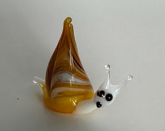 May include: A small glass snail figurine featuring a translucent white body with two antennae and dark eyes. The shell is crafted from swirled amber, brown, and white glass, creating a marbled effect that rises to a pointed peak at the top.