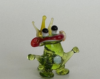 May include: A green glass figurine of a frog wearing a yellow crown and a red mouth. The frog has a blue glass eye and is standing on a white surface.